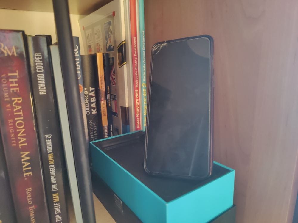 An older smartphone with a partially cracked display standing on a box on a bookshelf, surrounded by books — used as a secondary, rarely powered-on device.