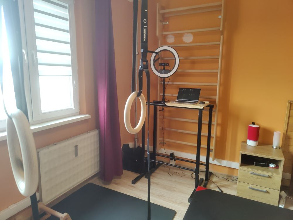 A small room with a compact training and work setup, including wall bars, gymnastic rings, a standing desk with a laptop and ring light, and a bed nearby, showing an integrated living and training environment.